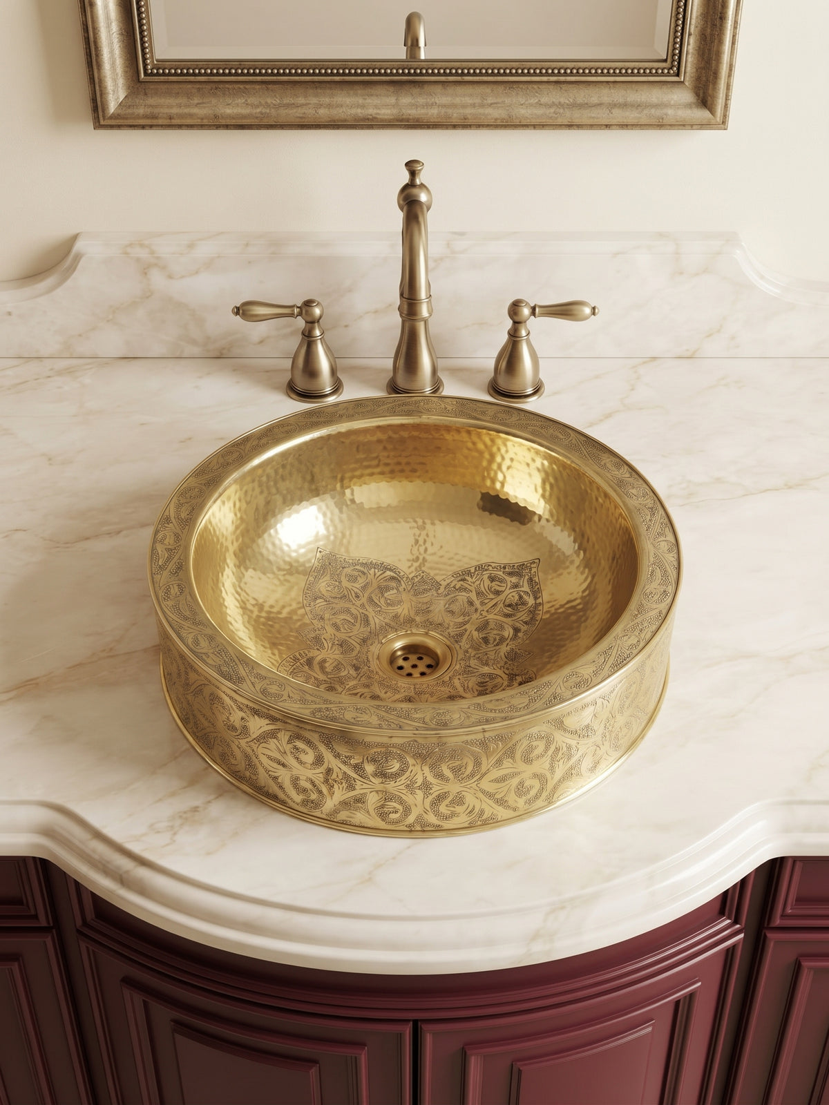 Round brass vessel sink with floral engravings and hammered interior on a cream marble vanity with brass faucet and burgundy cabinets.