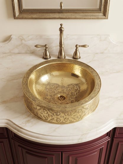 Round brass vessel sink with floral engravings and hammered interior on a cream marble vanity with brass faucet and burgundy cabinets.