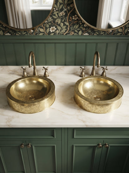 Two engraved brass vessel sinks on a white marble double vanity with green cabinets, brass faucets, and floral wallpaper.