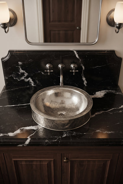 Round engraved silver vessel sink on a black marble vanity beneath a matching wall-mounted faucet, silver-framed mirror, and sconces.