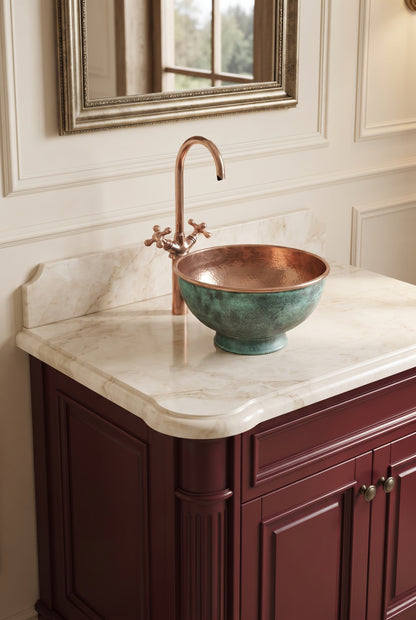 Green-patina copper vessel sink on a cream marble countertop above burgundy cabinetry, with a tall copper faucet and framed mirror.