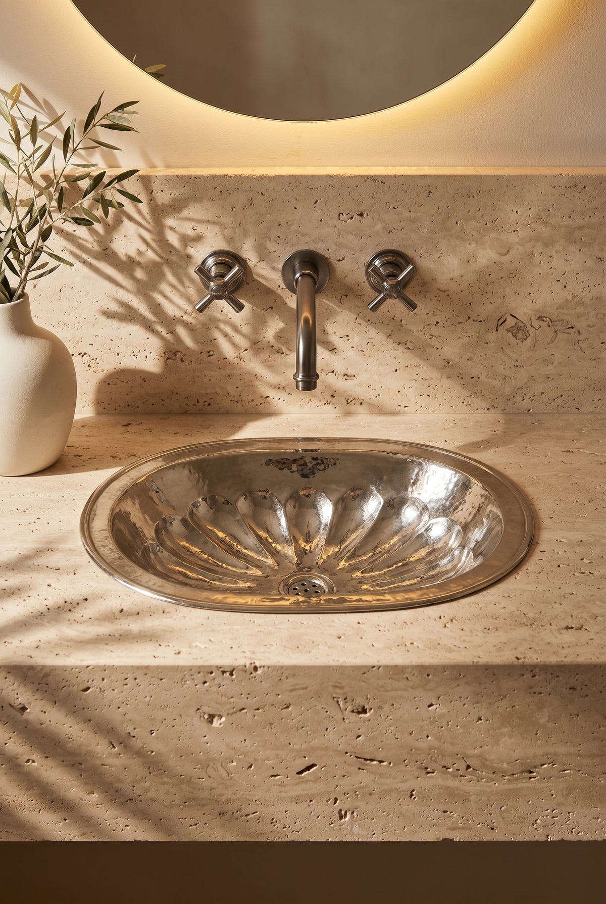 Oval silver sink with hammered flower-petal detailing in a travertine vanity beneath a wall-mounted faucet and round backlit mirror.