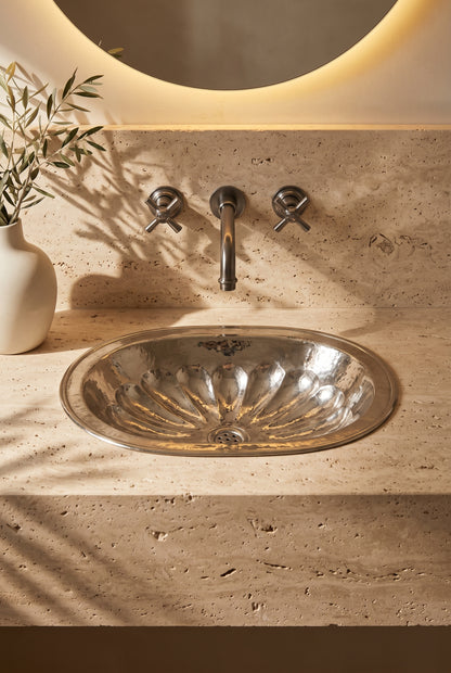 Oval silver sink with hammered flower-petal detailing in a travertine vanity beneath a wall-mounted faucet and round backlit mirror.