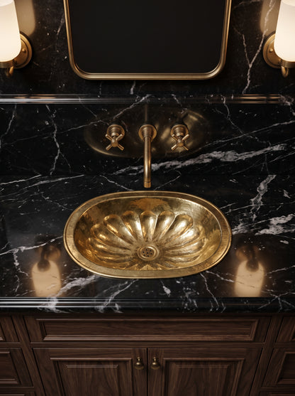 Oval brass sink with hammered flower-petal detailing in a black marble vanity with a brass wall-mounted faucet and dark wood cabinets.