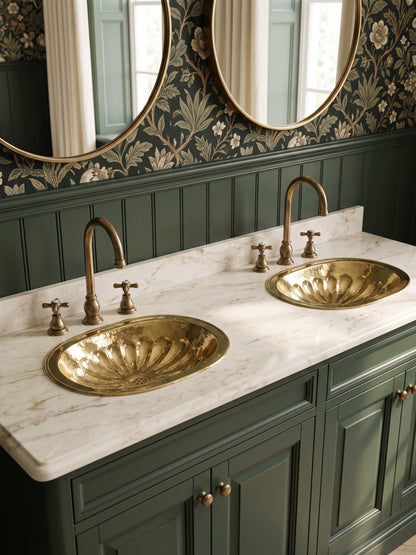 Two oval brass sinks with flower-petal detailing in a white marble double vanity with green cabinets, floral wallpaper, and oval mirrors.