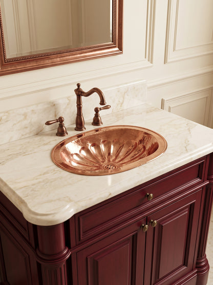 Oval copper sink with hammered flower-petal detailing in a cream marble vanity with matching faucet, burgundy cabinets, and copper-framed mirror.