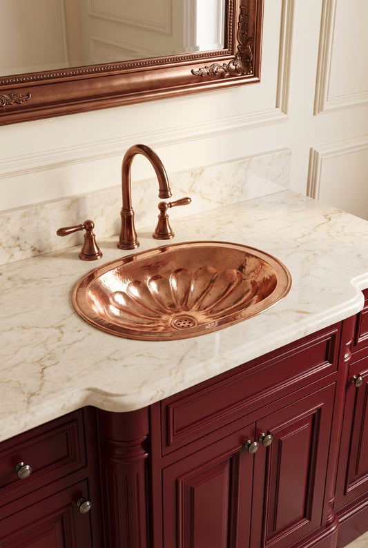 Oval copper drop-in sink with hammered flower-petal detailing in a cream marble vanity with matching faucet and burgundy cabinets.