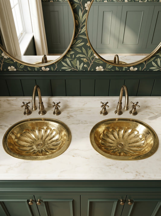 Two oval brass drop-in sinks with flower-petal detailing in a white marble double vanity with green cabinets, floral wallpaper, and oval mirrors.