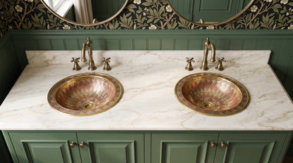 Two geometric brass sinks with copper and silver accents in a white marble double vanity with green cabinets, brass faucets, and oval mirrors.
