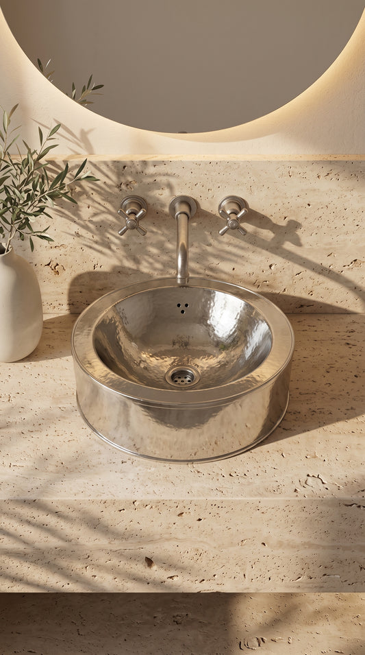Round hammered silver vessel sink on a travertine vanity beneath a wall-mounted faucet and round backlit mirror, beside a vase of leafy branches.