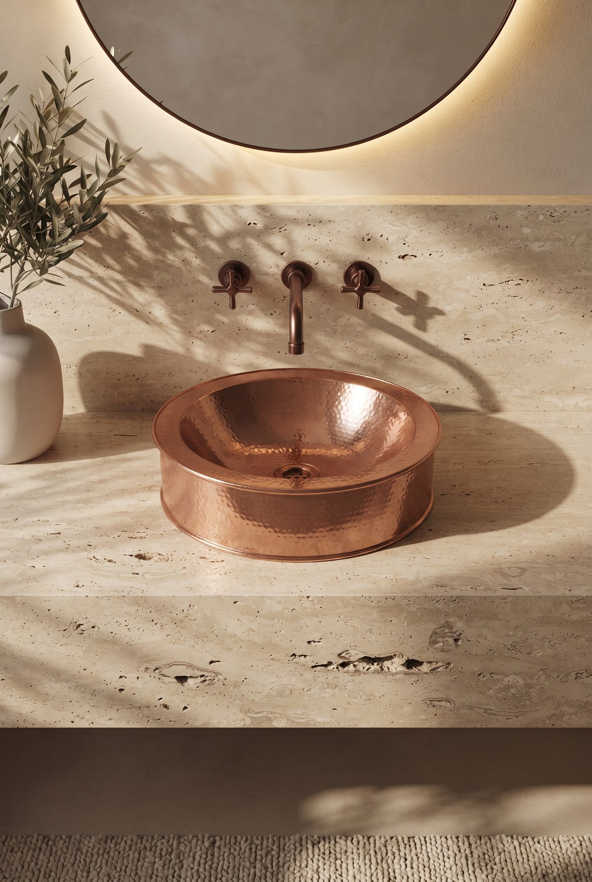 Round hammered copper vessel sink on a travertine vanity beneath a matching wall-mounted faucet and round backlit mirror.