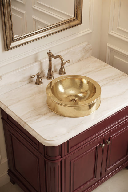Round hammered brass vessel sink on a cream marble vanity with brass faucet, burgundy cabinets, and framed mirror.