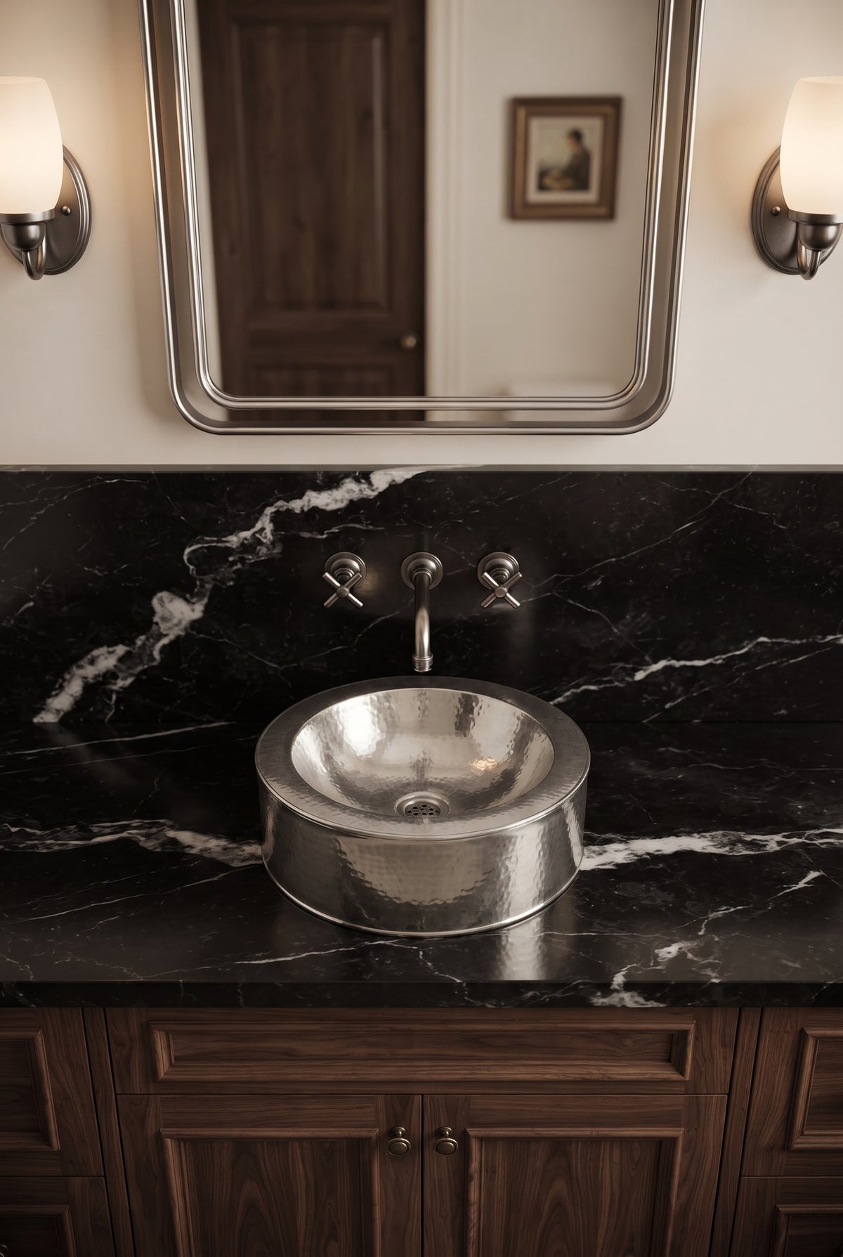 Round hammered silver vessel sink on a black marble vanity beneath a matching wall-mounted faucet, silver-framed mirror, and sconces.