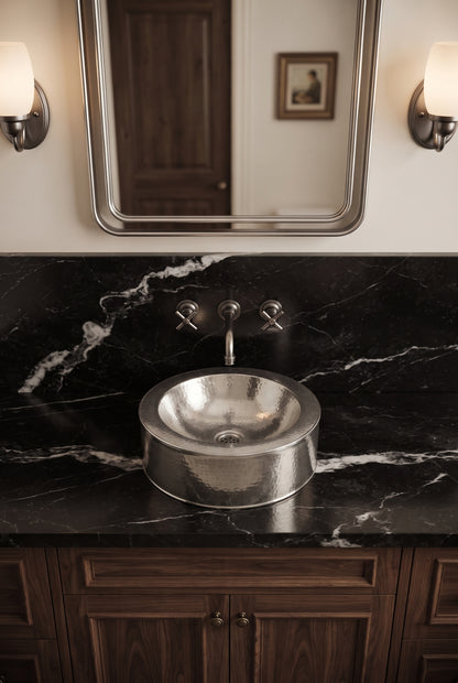 Round hammered silver vessel sink on a black marble vanity beneath a matching wall-mounted faucet, silver-framed mirror, and sconces.