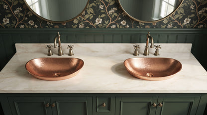 Two oval hammered copper vessel sinks on a cream marble countertop above dark green cabinets, beneath round mirrors and botanical wallpaper.