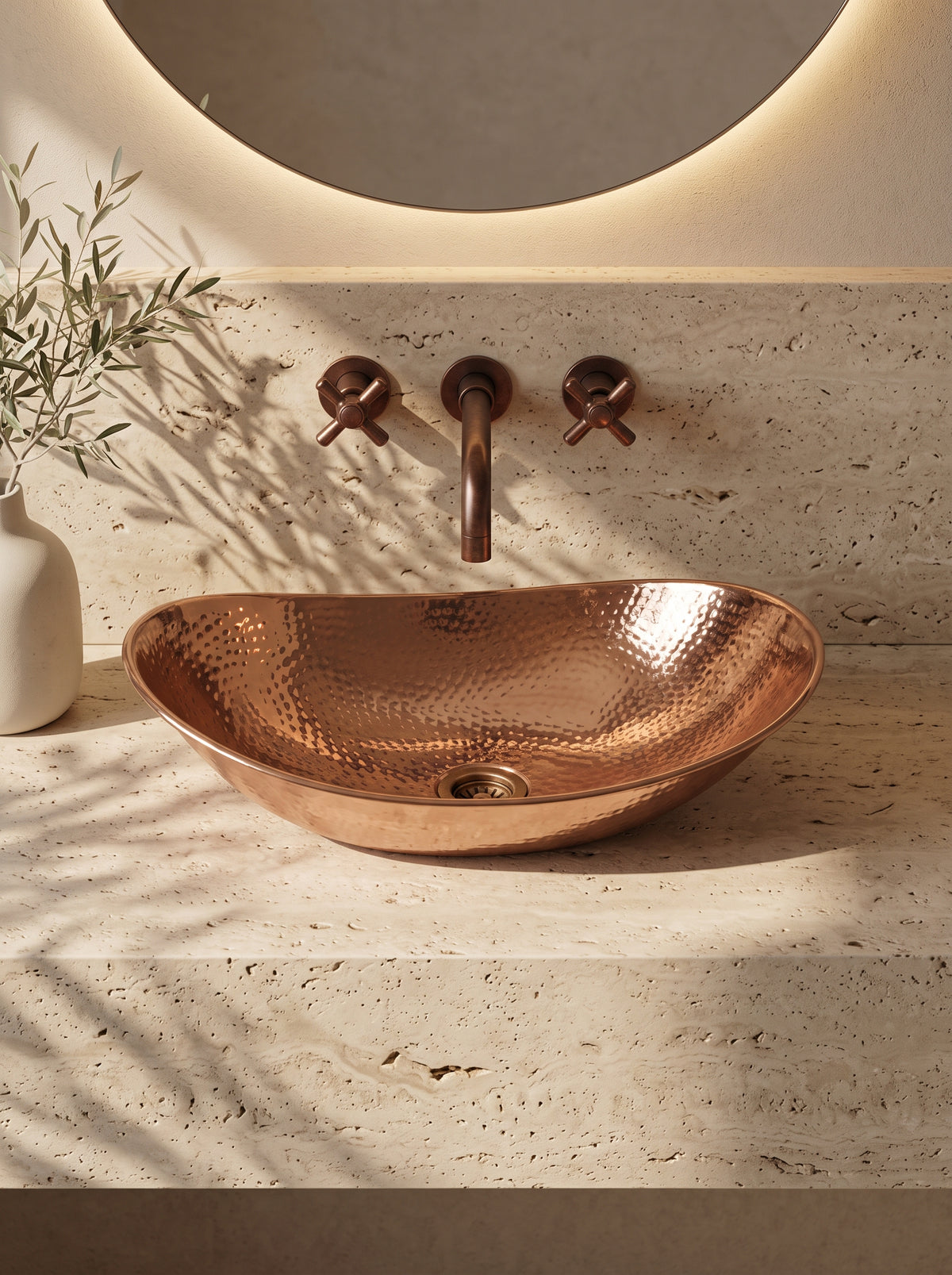 Oval hammered copper vessel sink on a beige stone vanity beneath a wall-mounted faucet and round mirror, beside leafy branches.