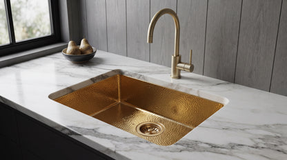 Hammered gold undermount kitchen sink with matching gooseneck faucet in a white marble countertop, beside gray wood paneling.