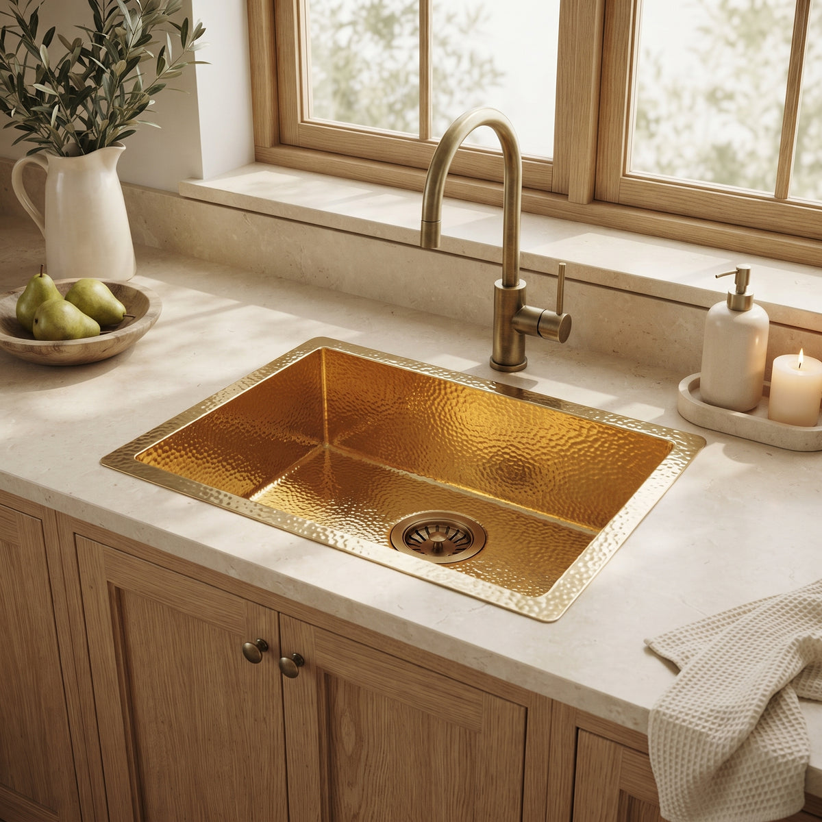 Hammered gold drop-in sink with matching faucet in a cream countertop above oak cabinets, styled with pears, soap, and a candle.