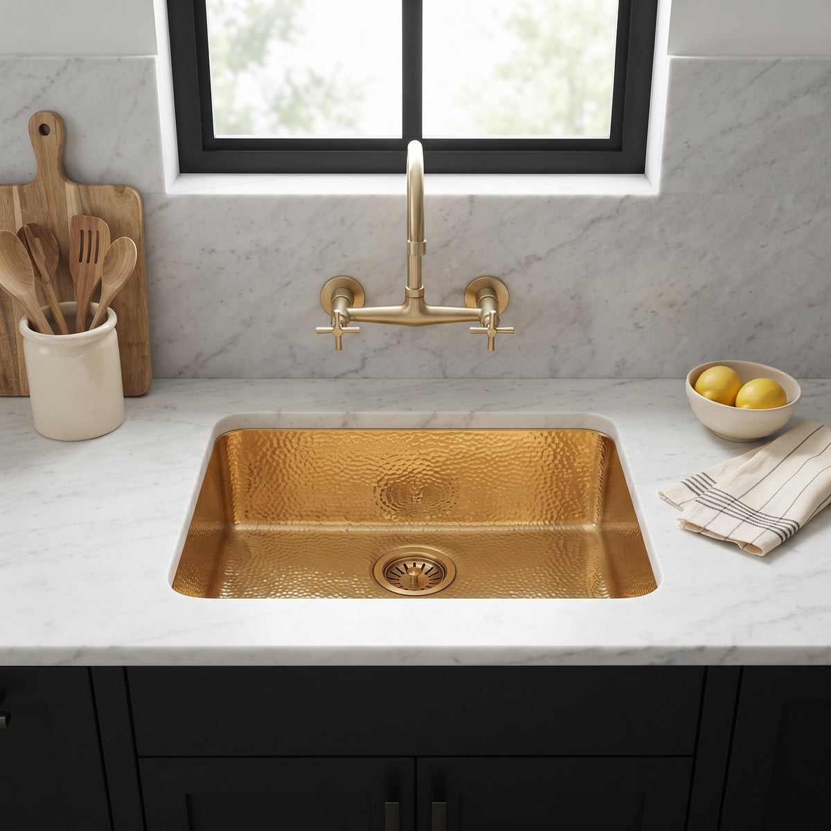 Hammered gold undermount sink beneath a wall-mounted brass faucet, with white marble surfaces, black cabinets, and a window above.