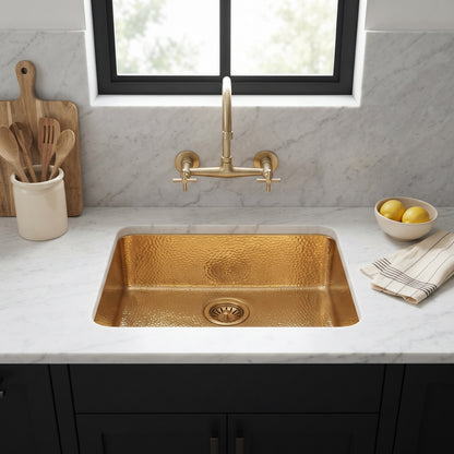 Hammered gold undermount sink beneath a wall-mounted brass faucet, with white marble surfaces, black cabinets, and a window above.