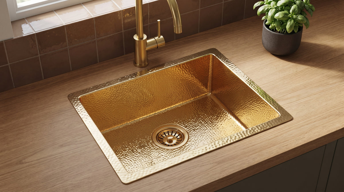 Hammered gold drop-in kitchen sink with a wide rim and matching faucet on an oak countertop, against brown tiles beside a basil plant.