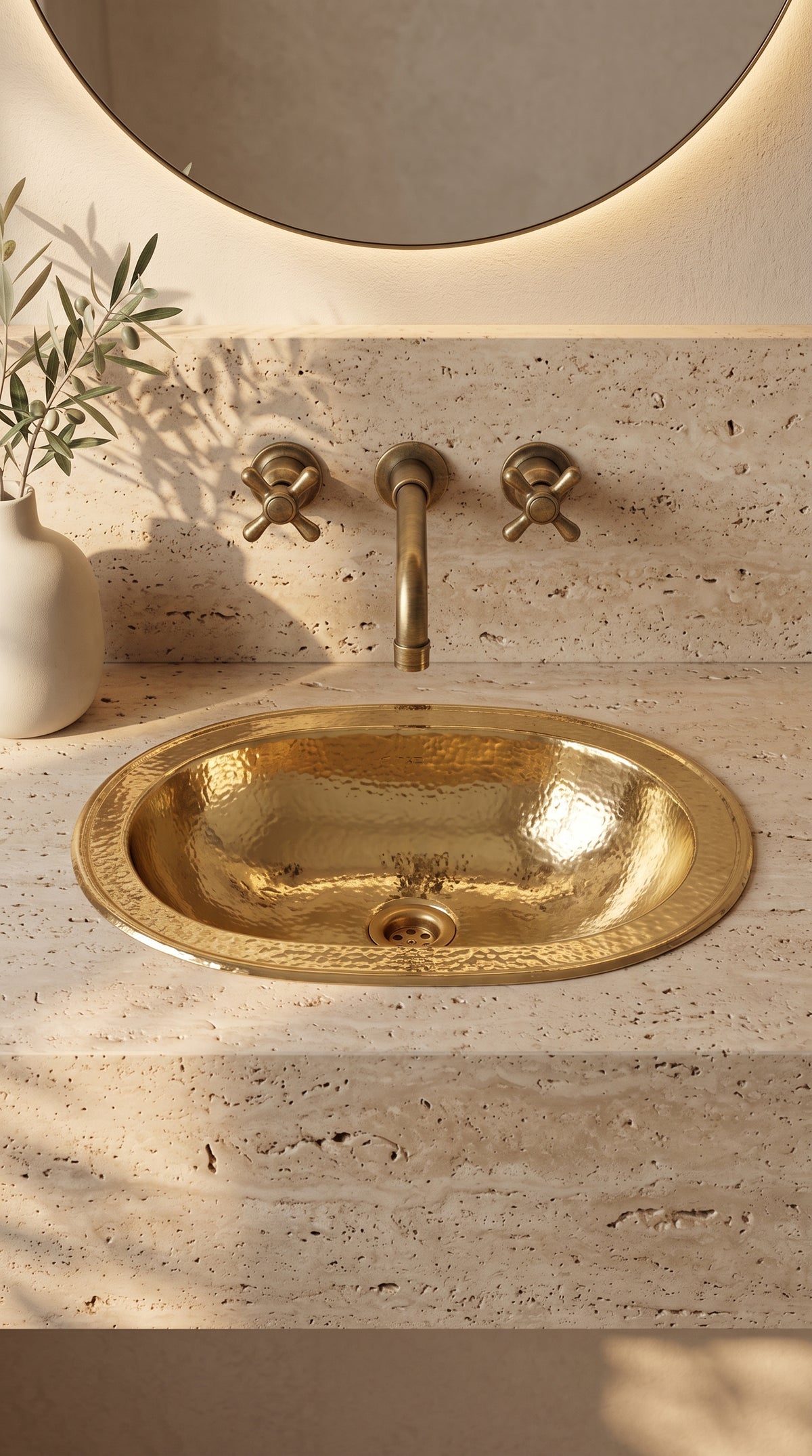 Oval hammered brass sink in a beige travertine vanity beneath a wall-mounted brass faucet and round backlit mirror, beside olive branches.