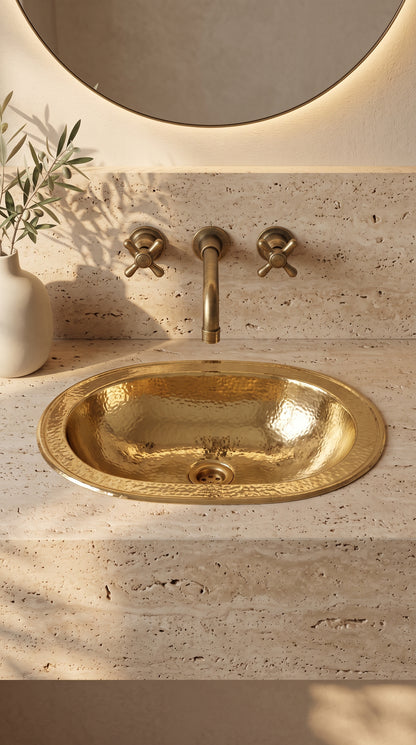 Oval hammered brass sink in a beige travertine vanity beneath a wall-mounted brass faucet and round backlit mirror, beside olive branches.