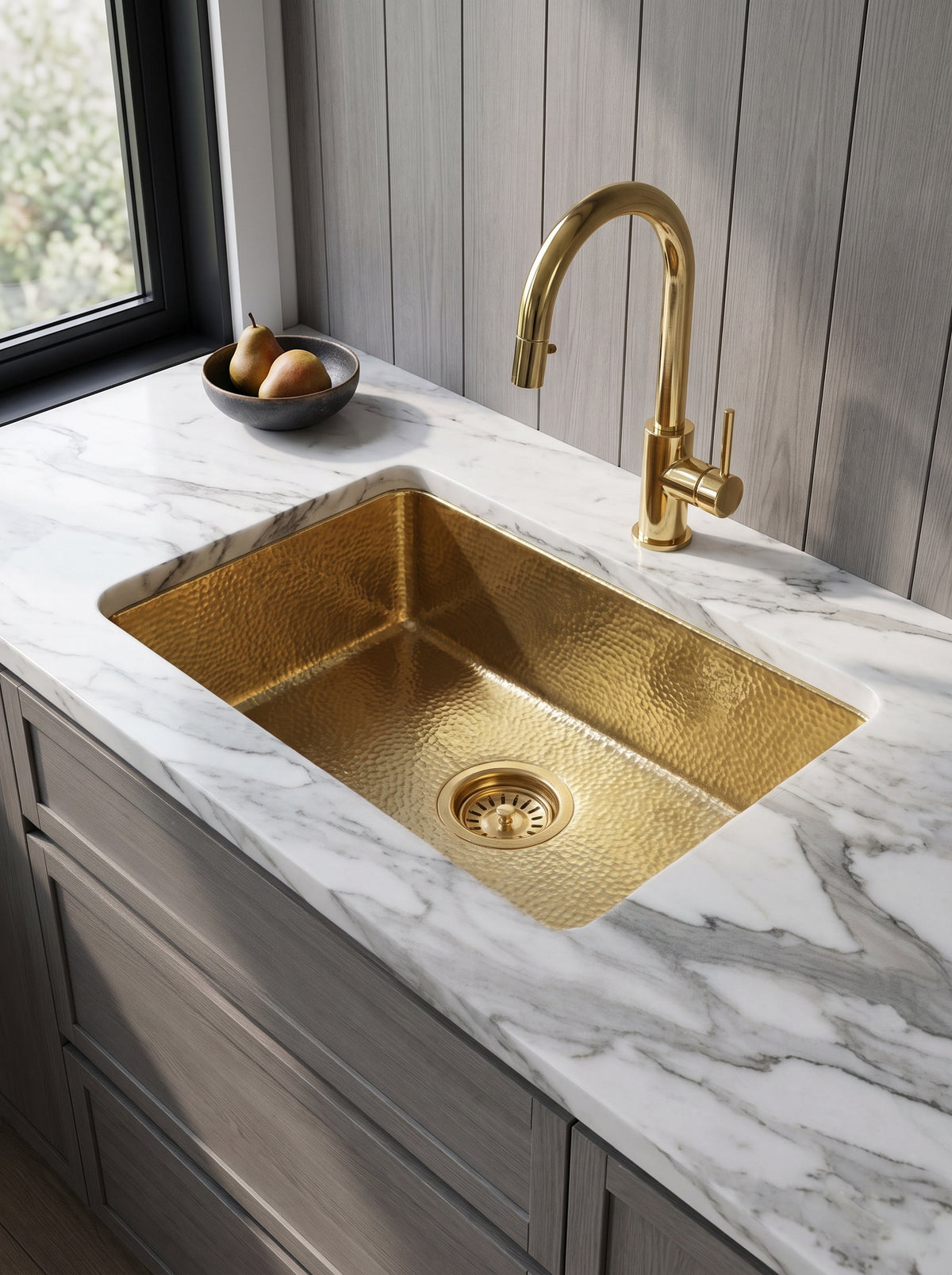 Rectangular hammered brass undermount sink in a white marble countertop, paired with a brass gooseneck faucet, gray cabinetry, and a bowl of pears.