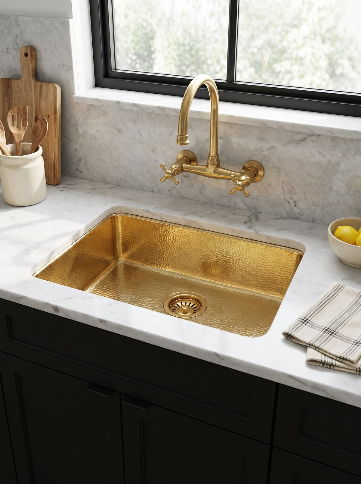 Hammered brass undermount sink beneath a wall-mounted brass faucet, surrounded by white marble, black cabinetry, and a sunlit window.
