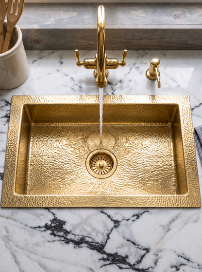 Overhead view of a rectangular hammered brass drop-in sink with water flowing from a brass faucet, set in a veined white marble countertop.