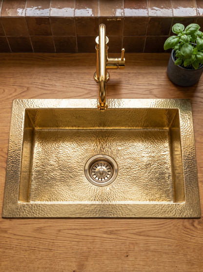 Rectangular hammered brass drop-in sink in an oak countertop, paired with a brass faucet, glossy brown tile backsplash, and potted basil.