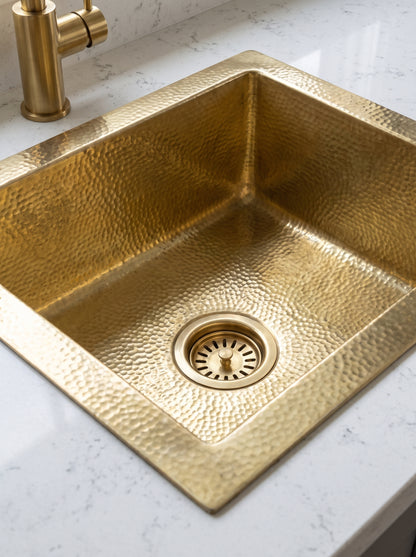Close-up of a hammered brass drop-in sink, highlighting its textured basin, exposed rim, rounded interior corners, and gold basket-strainer drain.