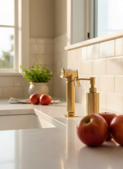 Unlacquered brass kitchen side sprayer and matching brass soap dispenser installed beside a white farmhouse sink on a bright quartz countertop.