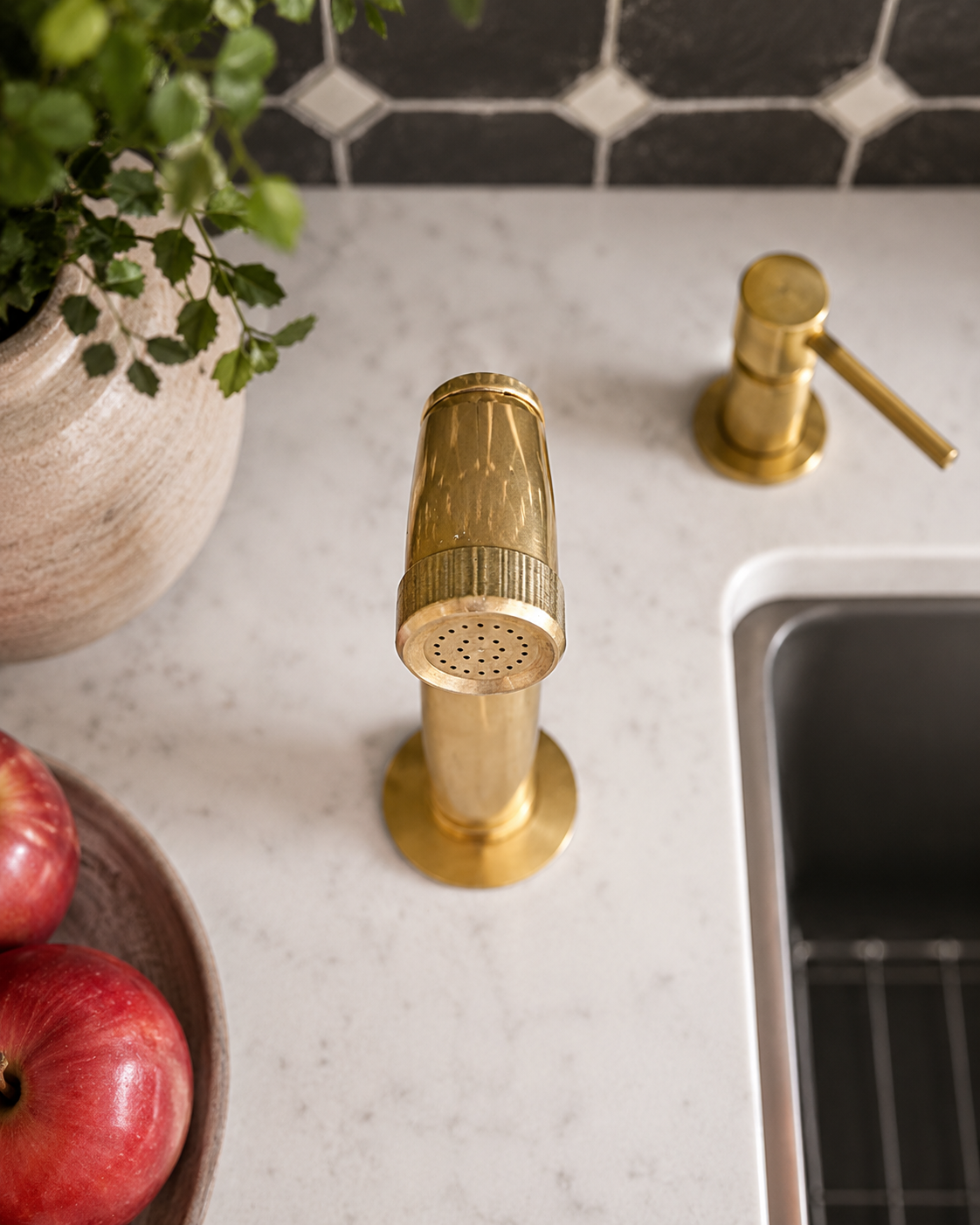 Top view of unlacquered brass kitchen side sprayer installed beside a stainless steel sink on a white marble countertop.