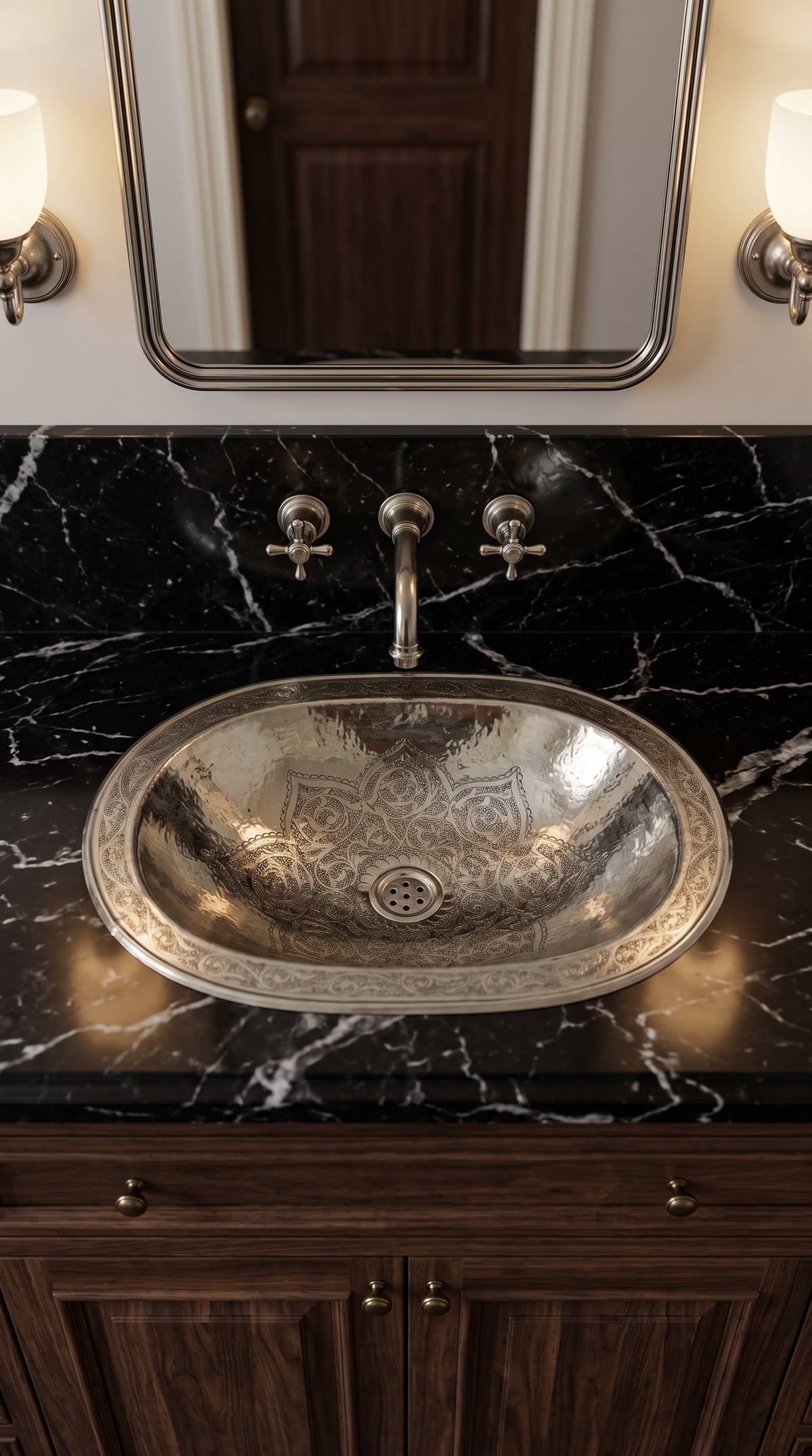 Oval silver sink with engraved floral motifs and decorative rim in a black marble vanity beneath a matching wall-mounted faucet and mirror.
