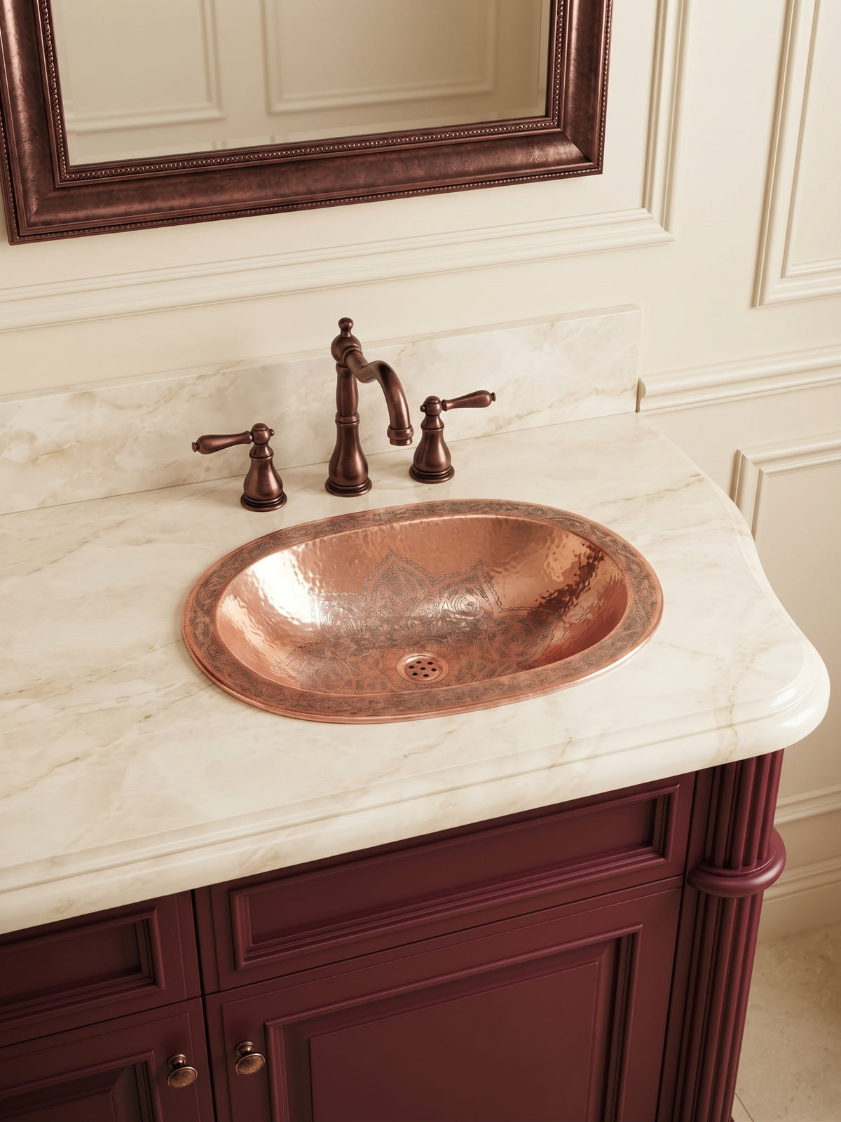 Oval hammered copper sink with engraved floral patterns set into a cream marble countertop, paired with a bronze faucet and burgundy vanity.