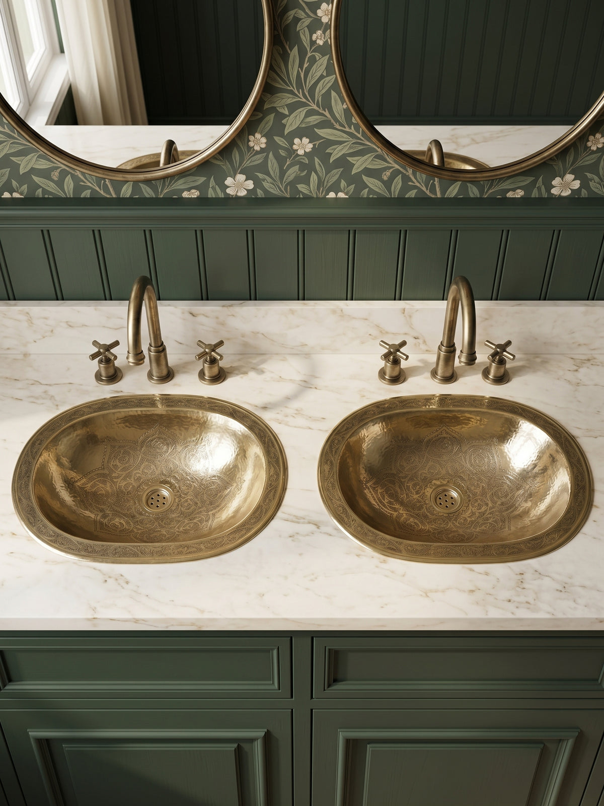 Two engraved oval brass sinks in a white marble double vanity with green cabinets, brass faucets, floral wallpaper, and oval mirrors.