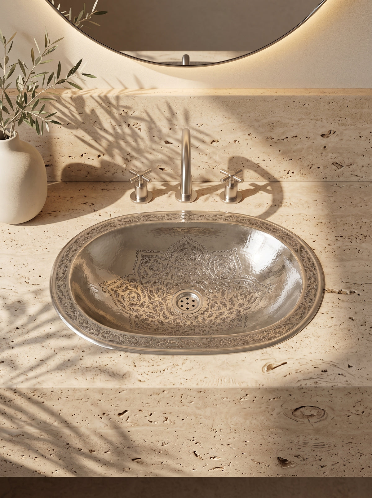 Oval silver sink with engraved floral motifs and decorative rim in a travertine vanity with matching faucet and round backlit mirror.