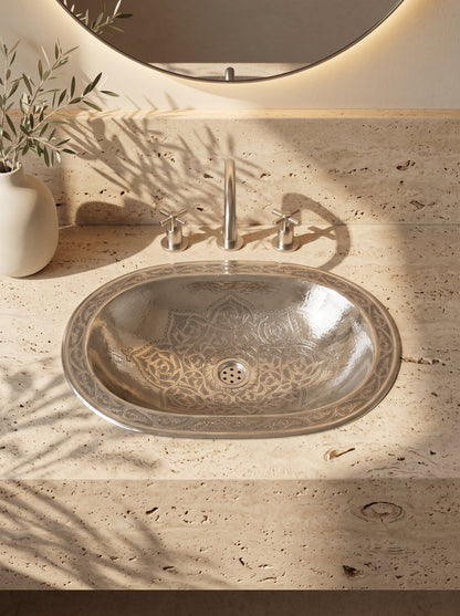 Oval silver sink with engraved floral motifs and decorative rim in a travertine vanity with matching faucet and round backlit mirror.