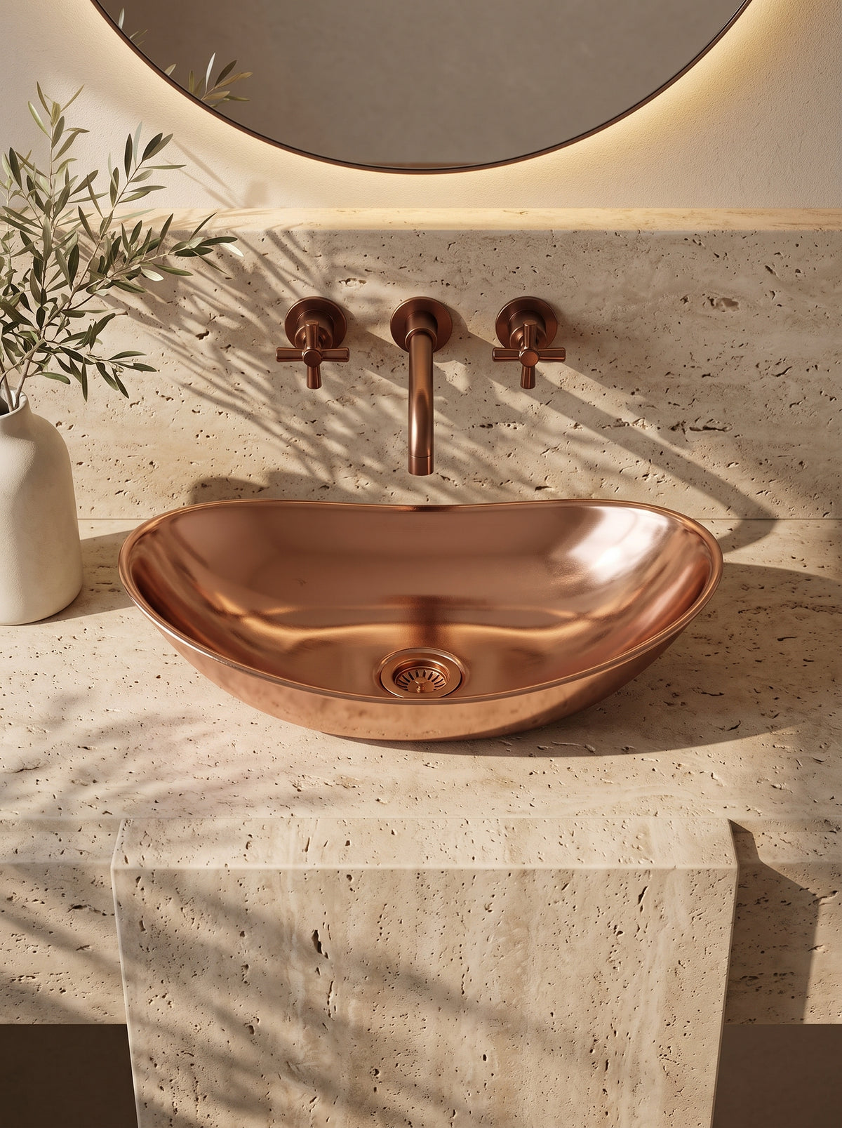 Oval copper vessel sink on a beige stone vanity beneath a wall-mounted copper faucet and round mirror, beside a vase of leafy branches.