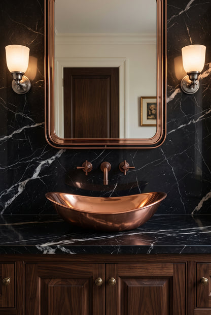 Oval copper vessel sink on black veined marble beneath a copper-framed mirror and two sconces, above dark wood cabinetry.