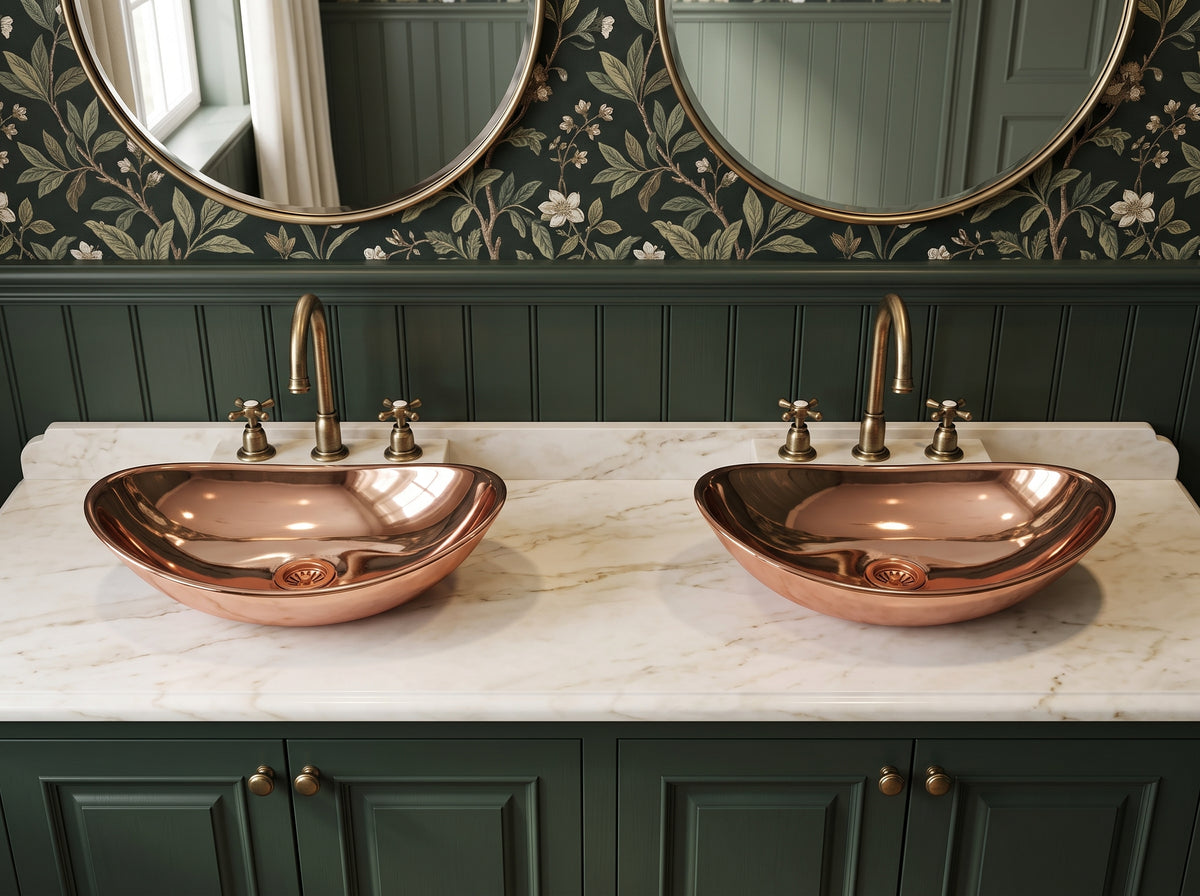 Two oval copper vessel sinks on a white marble countertop above dark green cabinets, paired with brass faucets, round mirrors, and botanical wallpaper.