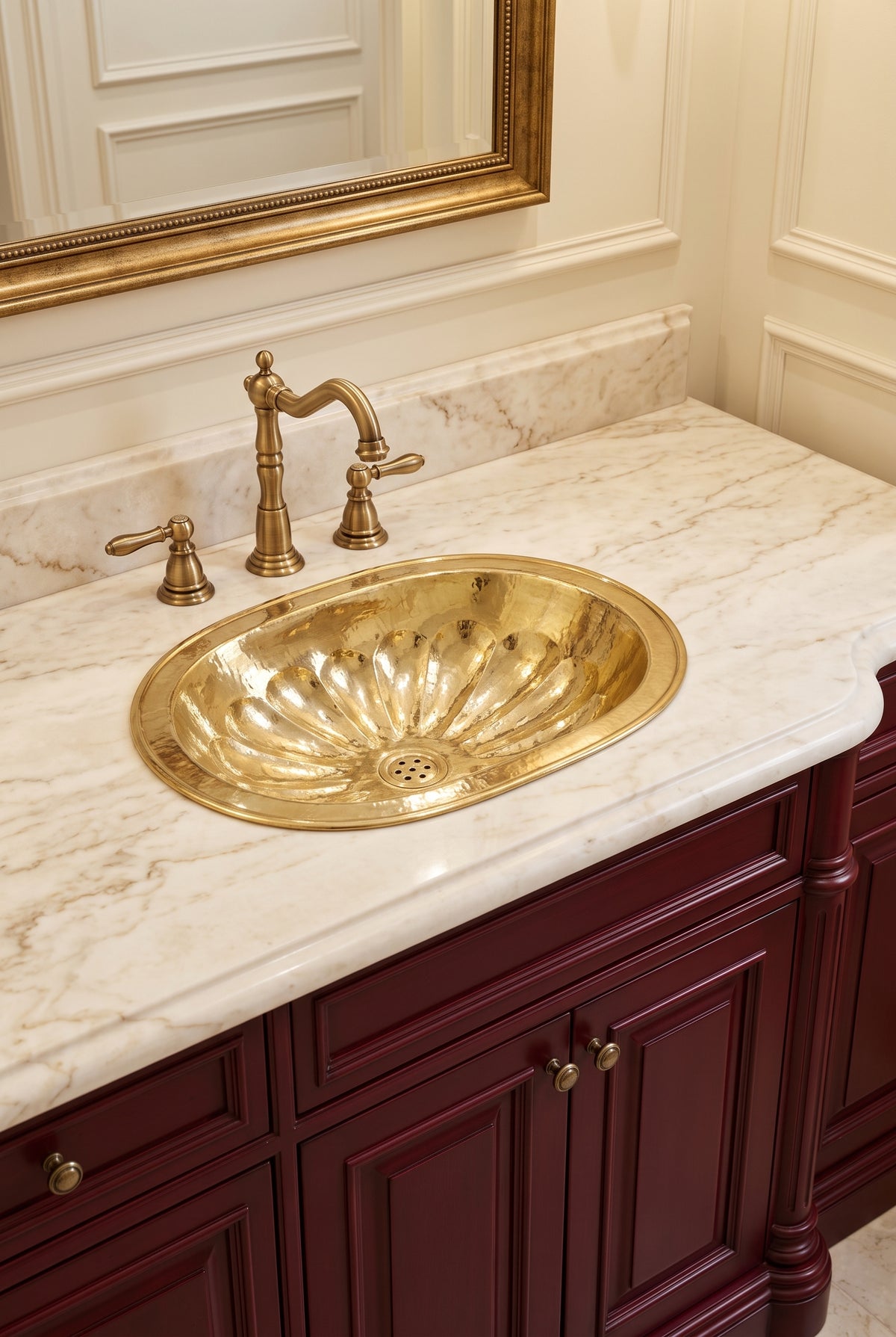 Oval brass sink with hammered flower-petal detailing in a cream marble vanity with matching faucet, burgundy cabinets, and gold-framed mirror.