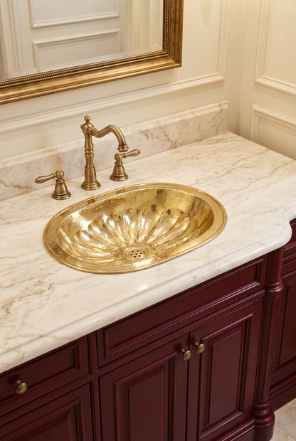 Oval brass sink with hammered flower-petal detailing in a cream marble vanity with matching faucet, burgundy cabinets, and gold-framed mirror.