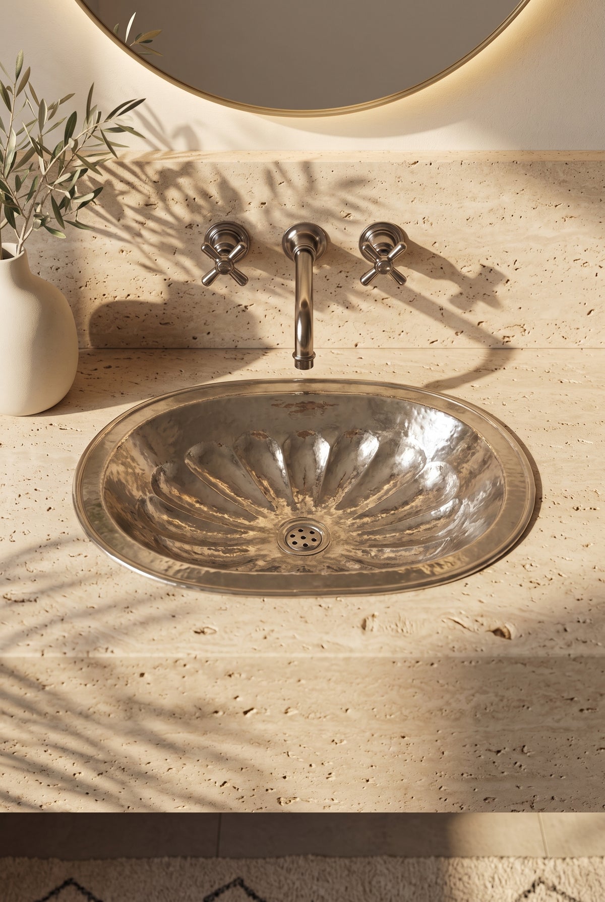 Oval silver sink with hammered flower-petal detailing in a travertine vanity beneath a wall-mounted faucet and round backlit mirror.