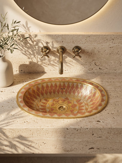 Oval sink with gold, copper, and silver geometric patterns in a beige travertine vanity beneath a brass wall-mounted faucet and round backlit mirror.