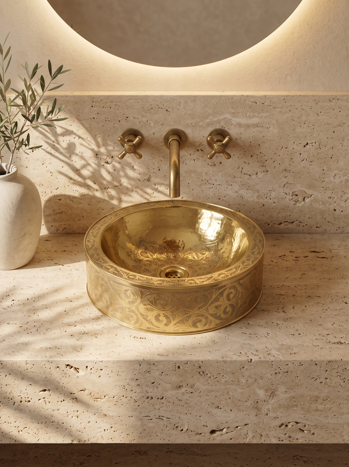 Round engraved brass vessel sink on a beige stone vanity beneath a wall-mounted brass faucet and round mirror.