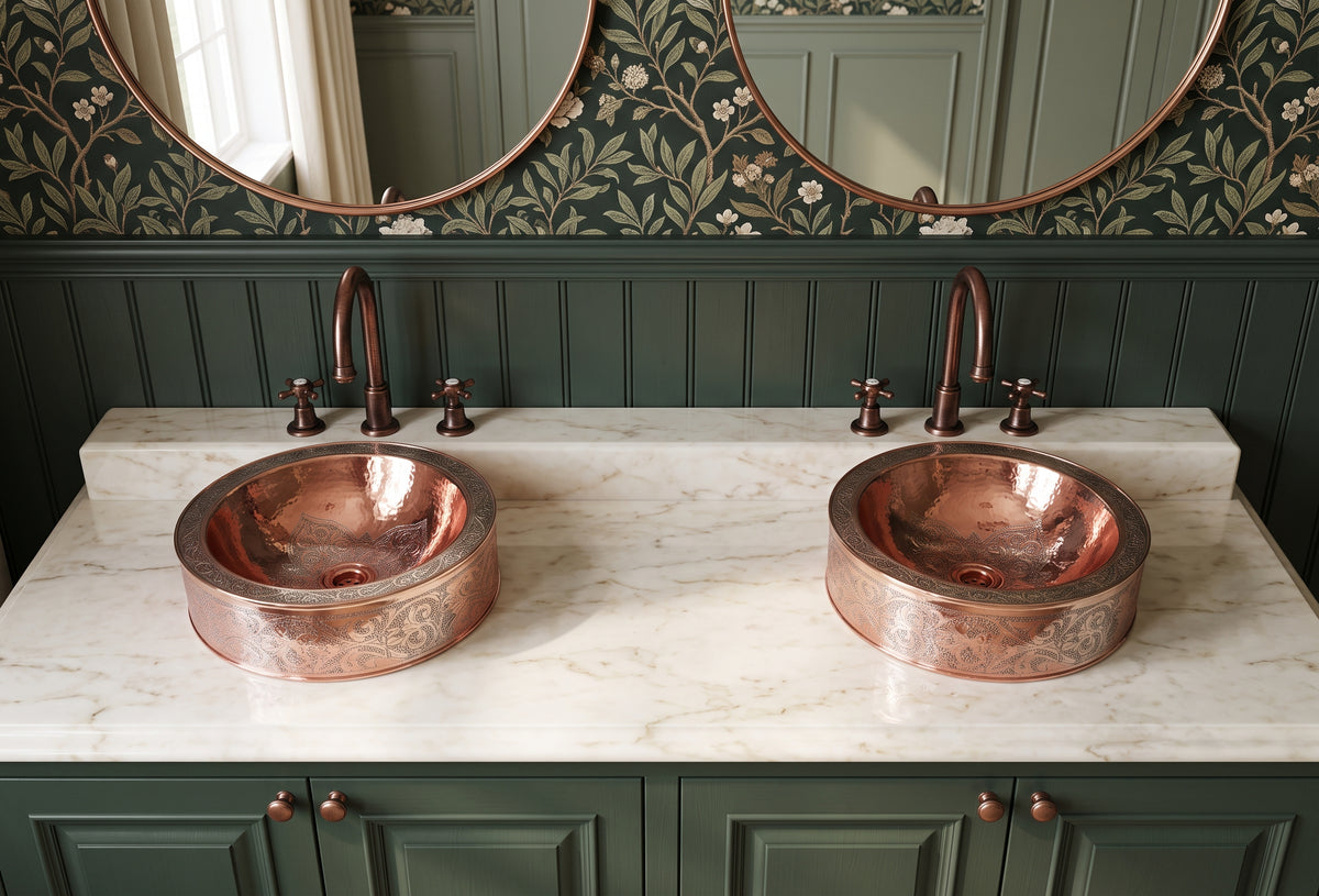 Two round engraved copper vessel sinks on cream marble above dark green cabinets, beneath round mirrors and botanical wallpaper.