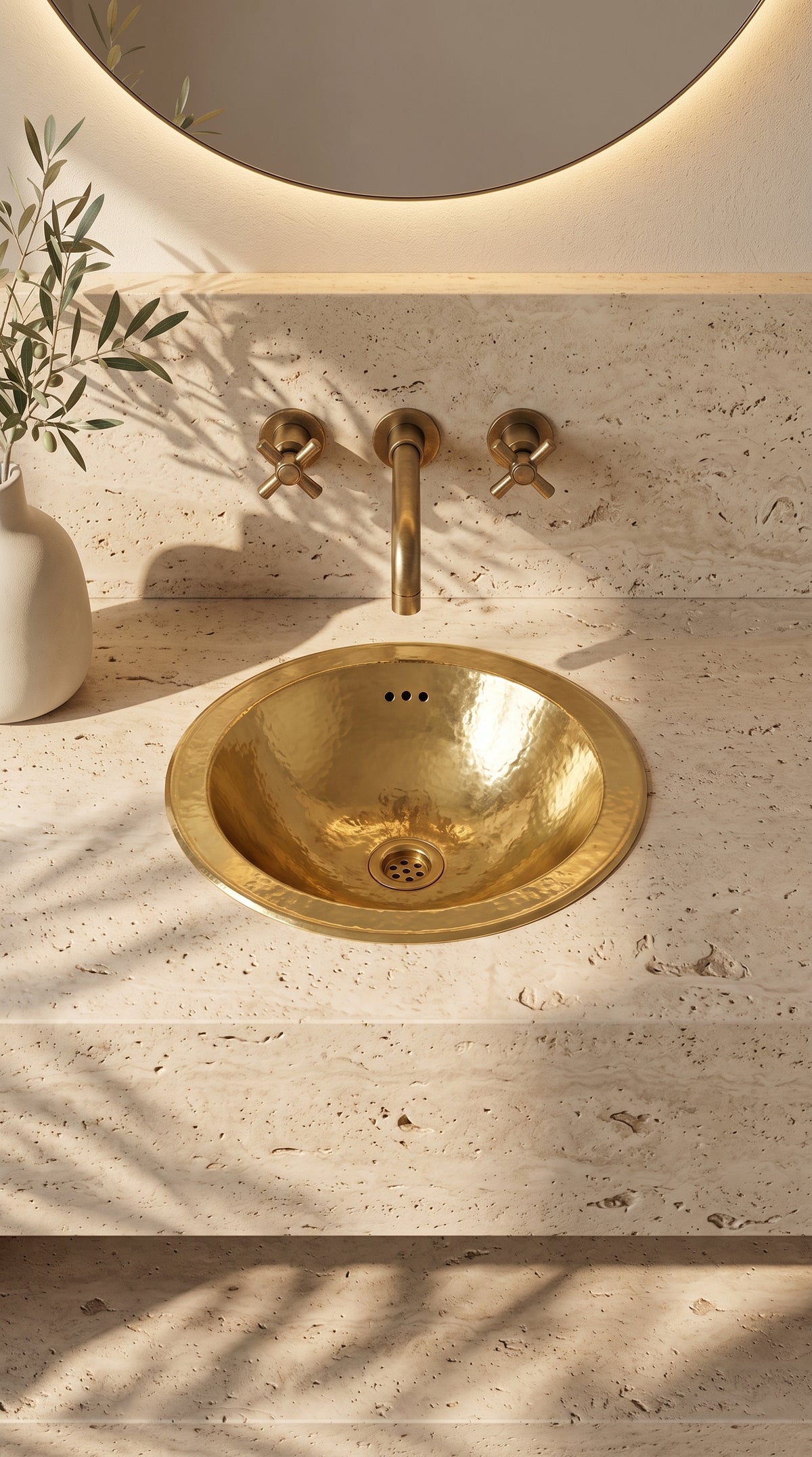 Round hammered brass sink in a travertine vanity beneath a wall-mounted brass faucet and round backlit mirror.