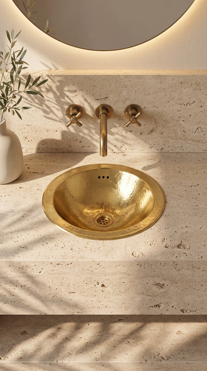 Round hammered brass sink in a travertine vanity beneath a wall-mounted brass faucet and round backlit mirror.